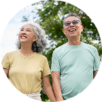Smiling senior East Asian couple holding hands and looking up while standing outdoors against a backdrop of green trees.